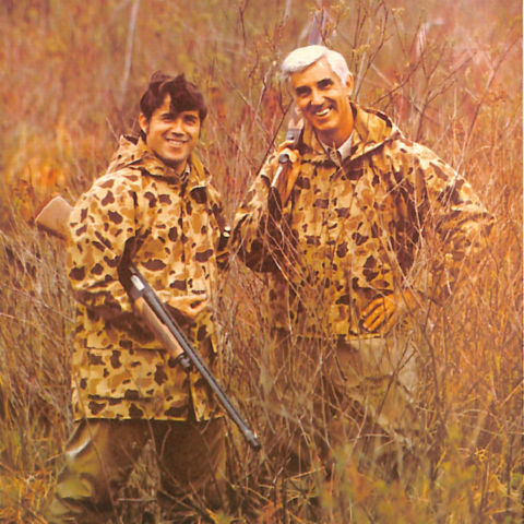 An image from the 1971 Orvis catalog of two hunters in a field wearing a camo-patterned jacket and holding a shotgun